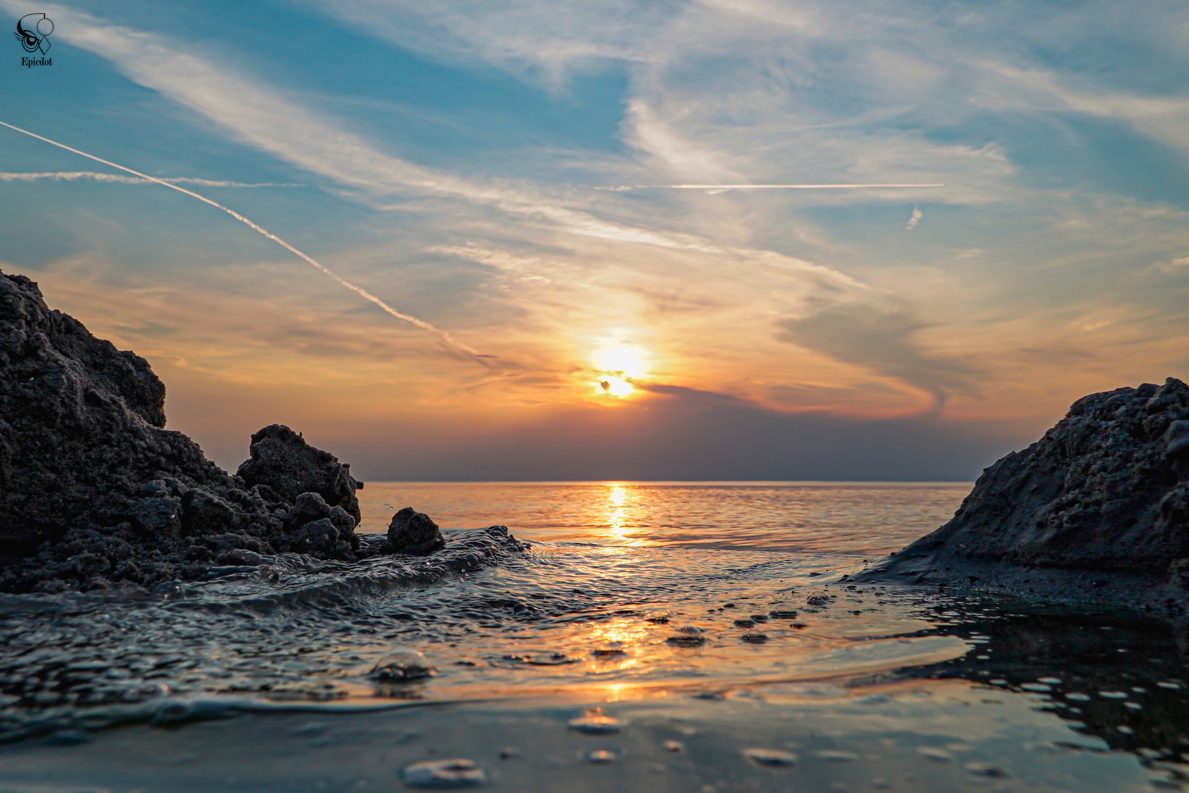 Sunset over calm sea waters with rocks in foreground and colorful cloud sky reflecting on the surface